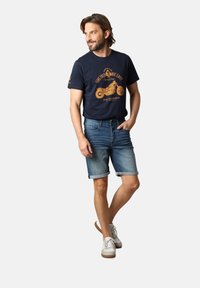 T-shirt graphique marine avec un design de moto orange, associé à un short en denim clair avec des ourlets retroussés. Des baskets blanches complètent la tenue.