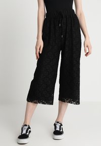 Čierne čipkované culottes s elastickým pásom, s jemným kvetinovým vzorom a skrátenou dĺžkou, v kombinácii s čiernymi teniskami.