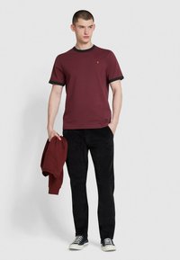 T-shirt bordeaux con collo a crew e rifiniture nere a coste, abbinata a pantaloni neri e sneakers blu, mentre tiene un sweatshirt bordeaux abbinato.