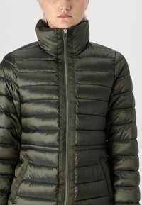 Polstret olivengrønn jakke med høy krage, horisontal quiltedesign, glidelås og sidelommer. Myk, blank stofftekstur.