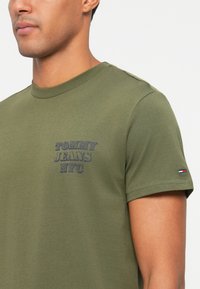 Vyras vilkintis žalios spalvos trumpomis rankovėmis marškinėliais su "Tommy Jeans NYC" tekstu ant krūtinės ir maža Tommy Hilfiger vėliavos logo ant rankovės.