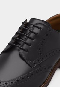Μαύρο δερμάτινο παπούτσι brogue με διακοσμητικές οπές και κλειστό κορδόνι σε καφέ σόλα, εμφανίζεται σε κοντινό πλάνο.