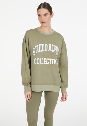 Frau trägt ein olivgrünes Sweatshirt mit dem Schriftzug "STUDIO AURE COLLECTIVE" und passende Leggings, steht vor einem weißen Hintergrund.