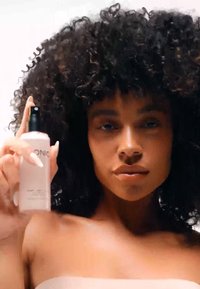 Bouteille spray transparente avec un embout noir, liquide rose à l'intérieur. Le modèle la tient avec confiance, avec des cheveux bouclés et volumineux et une peau aux tons neutres.
