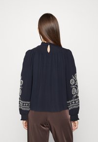 Marineblaue Bluse mit langen Ärmeln und floralem Stickdesign auf den Ärmeln. Schlüsselöffnungsdetail am Rücken. Weicher, fließender Stoff.