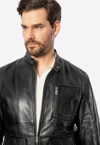 Chaqueta de cuero negra con cuello alto, cierre con cremallera en la parte frontal y bolsillo en el pecho. Textura suave con un ligero brillo y corte ajustado.