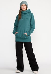 Jeune personne portant un sweat à capuche teal, un béret gris, un pantalon large noir et des baskets blanches, se tenant avec les mains dans les poches.
