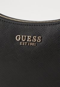Borsa nera a trama con logo dorato "GUESS" e dettaglio "EST 1981". La chiusura con zip accentua il design, mettendo in risalto un'estetica elegante e moderna.