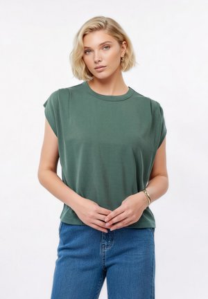 Kobieta z krótkimi blond włosami, ubrana w zielony top bez rękawów i niebieskie dżinsy, stojąca z rękami złożonymi przed sobą.