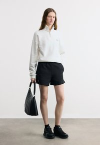 Γυναίκα που φοράει λευκό μπλουζάκι με φερμουάρ μέχρι το στήθος, μαύρο σορτς, μαύρα αθλητικά παπούτσια και κρατάει μαύρη τσάντα, στέκεται μπροστά σε λευκό φόντο.