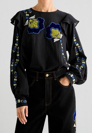 Zwarte blouse met lange mouwen met blauwe, groene en witte bloemrijke borduursels op de borst en mouwen, gecombineerd met een zwarte broek met vergelijkbare bloemrijke borduursels.