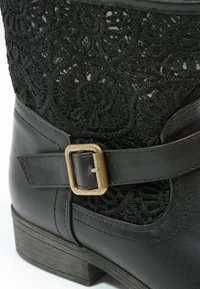 Botín negro hasta el tobillo con un panel lateral de encaje texturizado, parte superior de cuero y una correa curvada que presenta un detalle de hebilla dorada. Tacón bajo y grueso.