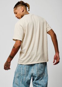 T-shirt texturé crème à manches courtes, coupe ample, vue arrière. Porté avec un jean en denim bleu délavé et usé. Tatouages notables sur les avant-bras.