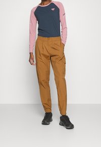 Long-sleeve shirt in navy blue with pink sleeves and logo, paired with tan trousers featuring a relaxed fit and side pockets, worn with black shoes.