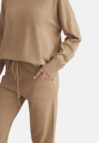 Ensemble de loungewear en tricot beige comprenant un pull à coupe ample avec des poignets côtelés et un pantalon assorti doté d'un cordon de serrage et de poches latérales.