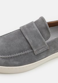 Chaussure à enfiler en suède gris avec un bout arrondi, dotée d'une large sangle sur le dessus et de coutures décoratives le long de la semelle.