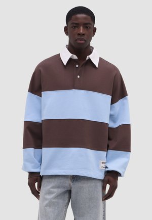 Junger Mann, der ein braun-hellblau gestreiftes Rugbyhemd mit weißem Kragen und hellen Jeans trägt, steht vor einem schlichten Hintergrund.