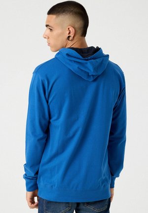 Sudadera con capucha azul hecha de tela suave, que presenta un corte holgado y mangas largas. El diseño incluye un bolsillo tipo canguro y puños de canalé.
