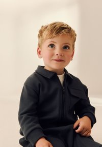 Combinaison zippée gris foncé pour enfants avec col, dotée d'une poche avant et de poignets côtelés. Tissu lisse avec une coupe décontractée.