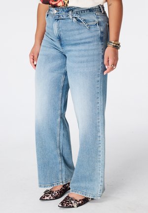 Personne portant un jean large bleu clair avec une ceinture nouée, des ballerines pointues imprimé léopard et plusieurs bracelets à un poignet.
