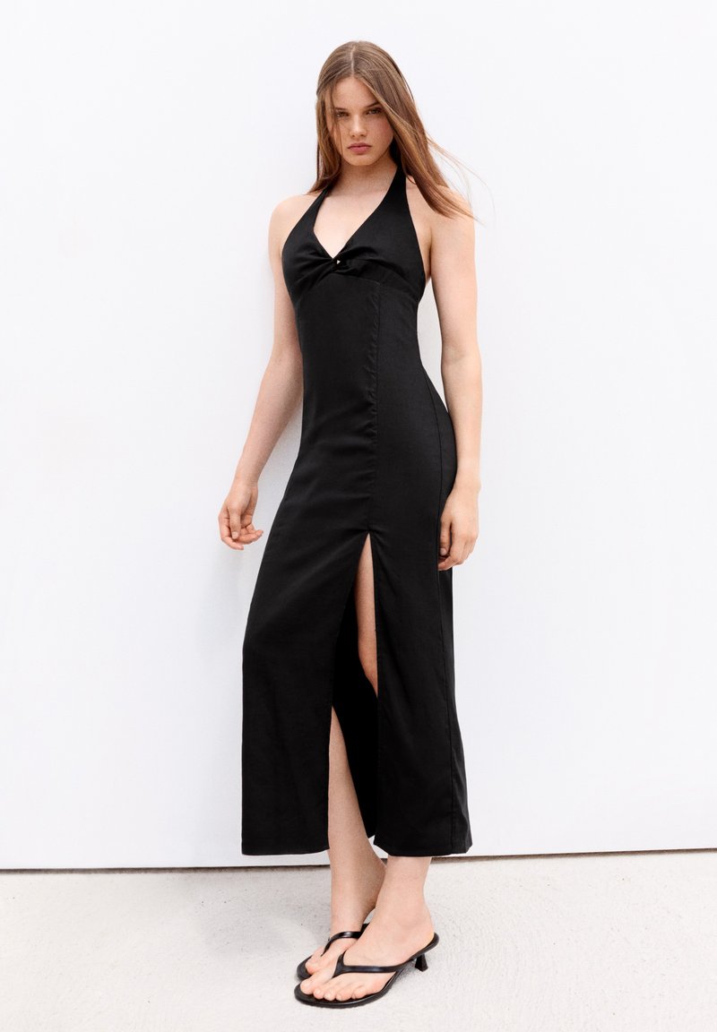 Jeune femme aux longs cheveux portant une robe noire dos nu avec une fente devant et des sandales à talons noires, debout contre un mur blanc.