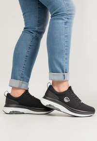 Schwarze Sportschuhe mit einer strukturierten Oberfläche, weißer Sohle und transparenter Luftdämpfung. Mit rundem Logo und Schnürdesign.