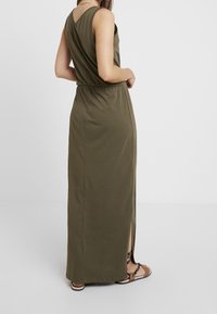 Oliwkowa długa sukienka z dekoltem w kształcie V z tyłu, elastycznym paskiem w talii i bocznymi rozcięciami. Wykonana z miękkiego materiału, charakteryzuje się minimalistycznymi detalami szycia.