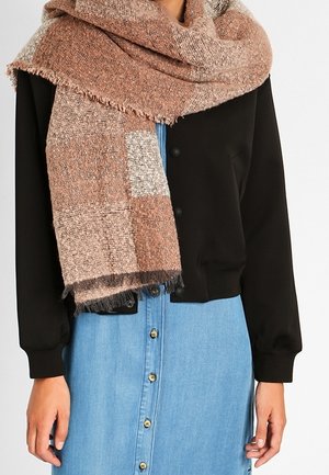 Femme portant une grande écharpe tricotée à carreaux rose et beige sur une veste noire et une robe en denim boutonnée bleue.