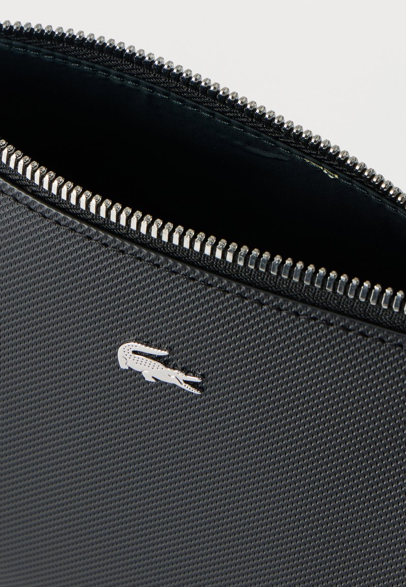Schwarzes, strukturiertes Täschchen mit teilweise geöffnetem silbernem Reißverschluss und kleinem silbernem Krokodil-Logo auf der Vorderseite.