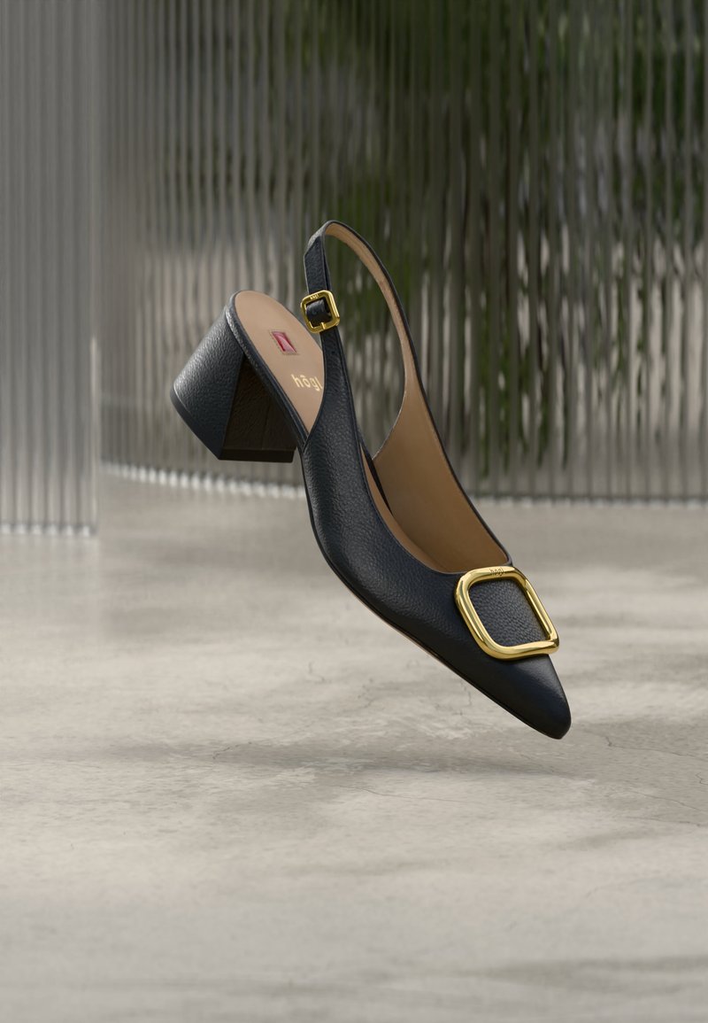 Μαύρο δερμάτινο γυναικείο παπούτσι τύπου slingback με χοντρό τακούνι και χρυσή αγκράφα, να αιωρείται πάνω από ένα λείο, ανοιχτό γκρι δάπεδο με φόντο υφή από γυαλί.