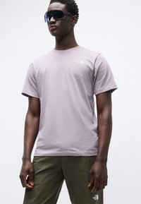 Ανοιχτό μωβ βαμβακερό T-shirt με στρογγυλή λαιμόκοψη και κοντά μανίκια, με μικρό κίτρινο λογότυπο στο στήθος. Φορεμένο με παντελόνι σε ελιάς χρώμα.