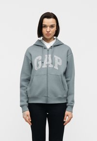 Femme aux cheveux courts et foncés portant un sweat à capuche à fermeture éclair bleu-gris avec le lettrage "GAP", une chemise blanche à col et un pantalon noir, debout devant un fond blanc.