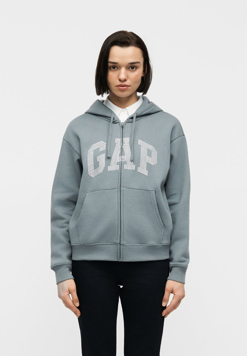 Femme aux cheveux courts et foncés portant un sweat à capuche à fermeture éclair bleu-gris avec le lettrage "GAP", une chemise blanche à col et un pantalon noir, debout devant un fond blanc.