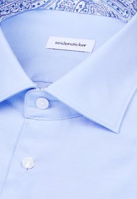 Chemise habillée bleu clair avec boutons blancs, étiquette de marque Seidensticker, et col intérieur à motif cachemire bleu et blanc.