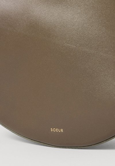Geantă rotundă din piele maro cu o textură netedă. Prezintă logo-ul embossat 'SOEUR' în auriu aproape de marginea de jos. Design minimalist.