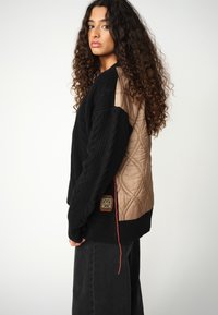 Donna con lunghi capelli ricci che indossa un maglione nero a trecce con schiena trapuntata beige e pantaloni neri larghi, vista di profilo.