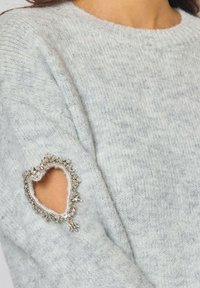 Pull en tricot gris clair avec une découpe en forme de cœur au niveau du coude, orné de détails en argent et d'une doublure intérieure douce.