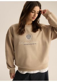 Beżowy, oversize'owy sweter z prążkowanymi mankietami. Zawiera haftowany napis "Różnorodne Dziedzictwo" oraz wzór naszywki. Nosi się go na białej koszuli.