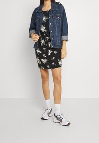 Jeansjack over een zwarte jurk met bloemenprint. Witte sportschoenen met accenten. Soepele texturen, aansluitende jurkvorm, korte mouwen.