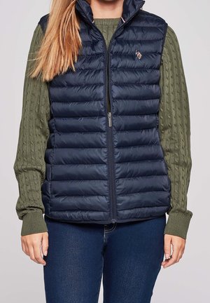 Vrouw die een marineblauwe doorgestikte mouwloze bodywarmer draagt over een olijfgroene kabelgebreide trui en donkerblauwe spijkerbroek, staand tegen een effen achtergrond.