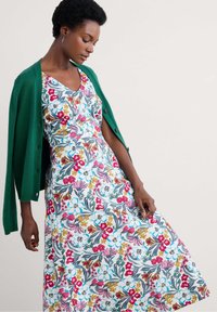 Robe fleurie avec un décolleté en V, ornée de motifs colorés de fleurs roses, rouges et jaunes sur un fond clair, accompagnée d'un cardigan vert.