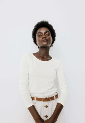 Jeune femme aux cheveux courts et bouclés portant une chemise blanche à manches longues et une jupe beige avec une ceinture marron, debout devant un fond clair uni.