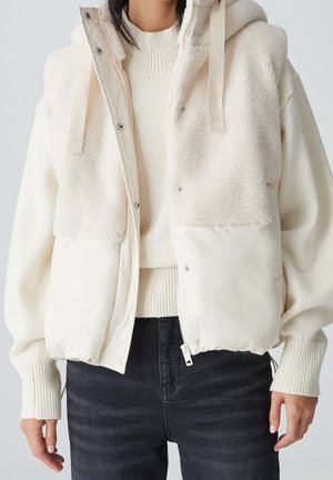 Femme portant une veste sans manches crème en sherpa et matelassée avec capuche à cordon, sur un pull en maille côtelée et un jean foncé.