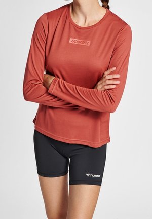 Kvinde iført rustfarvet langærmet top med "HUMMEL" logo og sorte Hummel-shorts, stående med armene over kors mod hvid baggrund.