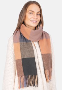Écharpe en tartan orange, marron et bleu marine avec des bords frangés. Matière douce qui se drape autour du cou, complétant un pull en tricot crème.