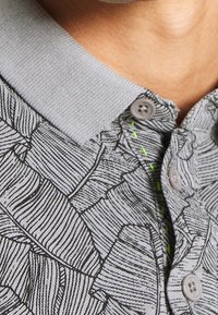 Polo gris avec un motif de feuilles texturé, doté d'un col gris clair et d'accents de couture verts. Les boutons sont de couleur gris moyen et ronds.