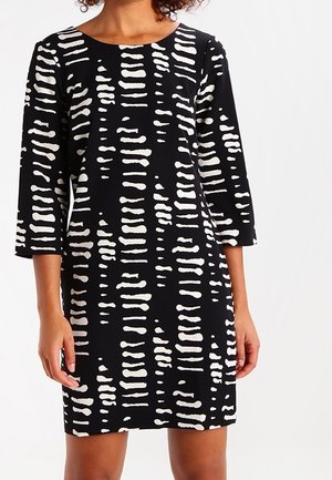 Mujer con un vestido negro con rayas horizontales irregulares blancas y mangas tres cuartos, de pie frente a un fondo liso.