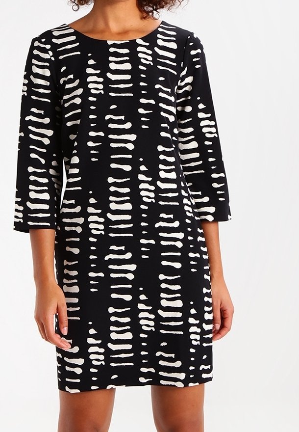 Femme portant une robe noire avec des rayures horizontales blanches irrégulières et des manches trois-quarts, debout devant un fond uni.