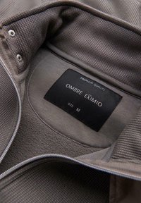 Gros plan sur le col d'une veste marron texturée avec une étiquette noire portant la mention "Qualité Premium Ombre Eximio Taille M".