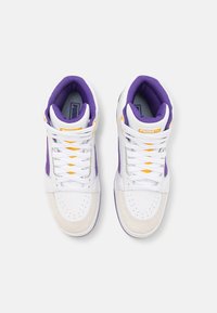 Paire de baskets montantes blanches avec des accents violets, des détails jaunes et des lacets blancs vues du dessus sur un fond uni.